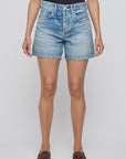 A person wearing a black sleeveless top, brown loafers, and Moussy’s Harrisburg Long Shorts—light blue high-waisted button fly jean shorts—stands against a plain white background.