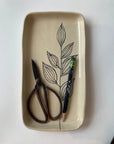 The Floral Small Tray by Faire, a rectangular hand-etched ceramic piece with a black leaf design, holds vintage scissors and a green-eraser pencil on a white surface, capturing subtle botanical symbolism.