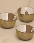 Three small Dansbury Snack Bowls by Faire, artisan crafted with textured metallic gold exteriors and smooth glossy white interiors, are arranged on a light beige surface.