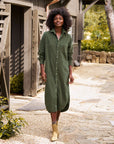 A woman with curly hair walks outdoors, smiling in the Frank & Eileen RORY Maxi Shirtdress and beige ankle boots. The rustic wooden building and greenery create a relaxed, stylish California-chic vibe.
