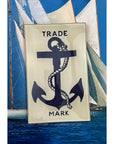 The Faire Shipman's Anchor Glass Tray showcases reverse decoupage artistry with a black anchor and rope labeled "TRADE MARK," displayed before a sailboat photo with white sails and blue sky.