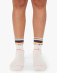 A person wearing "The Fold Please" cream-colored, ankle-length tube socks by Mother, with "Mother" and "Fucker" in pink embroidery, frilled hems, and blue, yellow, and orange striped cuffs stands on a white surface.
