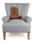 A light gray upholstered armchair with wooden legs and silver studs features the Faire Brown Cowboy Boot Pillow, a poly velvet throw pillow shaped like a brown cowboy boot, placed on the seat.