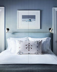 A bed with white linens and embroidered pillows evokes the style of Robert Stilin New Work by Hachette Book Group, centered in a blue-walled room between two doors, sunlight streaming through blinds onto reading lights and art above the bed.
