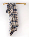 The Faire Cook's Cloth, a blue, beige, and white plaid cotton-linen blend kitchen towel, hangs on a brass towel bar against a plain white wall.