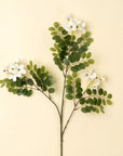 The Indaba Locust Flower Branch features green oval leaves and small white faux flowers, arranged as a decorative accent against a plain light beige background.