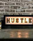 The Faire Lighted Sign featuring "HUSTLE" in bold black letters rests on a wooden surface by a brick wall, alongside stacked books and a white cup with saucer.