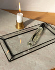 A lit white candle in a brass holder sits on a textured surface next to the Faire Glass Valet Tray, which holds a clear perfume bottle with a black cap. Warm light casts soft shadows across this elegant arrangement.