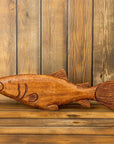 The Faire Rustic Carved Wood Fish Figure features intricate details and is displayed against a wooden plank wall, making it an ideal accent for rustic lake house or coastal cabin decor.