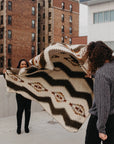 Tsachila Blanket in Chocolate, a large geometric boho woven throw held outdoors by two people.