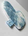 The Faire Heart Eye Pillow, featuring a heart shape and blue-and-white tie-dye cotton, rests on a white surface. A small blue fabric tab extends from the side—ideal for gentle anxiety relief.