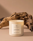 A white 14oz. "Cattleman" soy wax candle by Faire, known for its masculine scent, is placed in front of weathered driftwood on a beige surface with a neutral background.