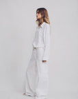 A woman stands against a plain light background, wearing a loose-fitting white button-up shirt and Denimist’s 100% cotton DECK PANT WHITE WASH. Her long hair is down, and she faces to the side with a relaxed posture.