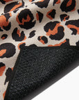 A close-up of the Faire Leopard Yoga Towel shows its brown, black, and orange leopard print. The flipped corner reveals a textured black, slip-resistant backing for secure grip.