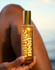 A hand holds a bottle of Faire Shimmer Body Oil—a nourishing, antioxidant-rich golden formula—against a backdrop featuring a person’s leg and an out-of-focus outdoor setting, possibly near water.