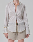 Wearing the camilia shirt S26 by Citizens Of Humanity/AGOLDE—a shiny beige striped cotton-silk button-down with flared cuffs—paired with beige shorts, a black belt, gold necklaces, and a bracelet against a light gray background.