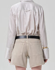 A person is seen from behind wearing the Citizens Of Humanity/AGOLDE camilia shirt S26, a beige striped cotton silk blend with oversized cuffs, tucked into light beige denim shorts and styled with a black belt and gold bangle.
