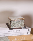 The Whisper Lidded Box by Accent Decor, a square textured ceramic piece, rests atop "THIS IS HOME" and "BOHÉMIANS," providing stylish jewelry storage and serving as a chic decorative accent on a wooden surface with a neutral backdrop.