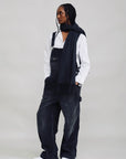 Black corduroy wide-leg SLOUCH BIB OVERALL by Denimist worn over a white shirt with a matching scarf.