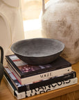The Rossetti Bowl SM by Accent Decor in matte black rests on a stack of art and fashion books, including Vogue, beside a large textured white vase on a wooden surface in a bright, stylish interior.