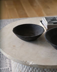 The Rossetti Bowl SM by Accent Decor in matte black sits on a round, light stone table beside books, with another handcrafted terracotta bowl and a patterned rug visible in the background.