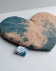 The Faire Heart Eye Pillow, featuring a blue and peach tie-dye cotton design, rests on a white surface. This heart-shaped pouch is perfect for anxiety relief and includes a small tag at the bottom edge.