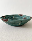 The Birava Large Bowl by Faire is a shallow, round woven basket in teal, brown, cream, and blue—ideal as handmade decor or Rwanda wall art. Shown on a light textured surface with a plain white background.