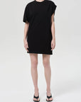 A person stands against a plain background wearing the Citizens Of Humanity/AGOLDE Roma Dress, a loose-fitting black organic cotton mini dress with an asymmetrical drape on one shoulder and short sleeves, paired with black open-toe heels. Face not visible.