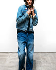 A person stands indoors against a plain white wall, wearing a blue denim jacket with a shearling collar, an Indigo French Sailor Pant by Free City (sparrow, LLC), a black shirt, and black shoes, with hands holding the jacket collar.