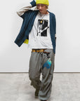 A person wearing a yellow beanie, Free City Cashmere Lux Cardigan, graphic white T-shirt, loose gray pants with blue text, and green sneakers stands indoors on a concrete floor, looking down with one hand on their head.