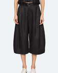 A person stands against a plain light background wearing Sea New York's Angelina Suiting Culotte—loose, wide-leg black polyester pants with a belt—paired with a black top and white shoes.
