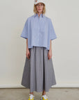 A person stands against a plain background wearing a light blue oversized button-up shirt, yellow sneakers, a white baseball cap with colorful patches, and the Le Cargo Skirt by Aquarius Cocktail—a long gray pleated cargo skirt with an elastic waist.