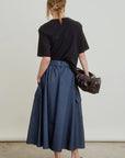 A woman with blonde hair in a low, messy bun wears a black t-shirt, Aquarius Cocktail's Le Cargo Skirt in Japanese cotton poplin denim with oversized pockets and an elastic waistband, black slip-on shoes, and holds a dark brown bag.