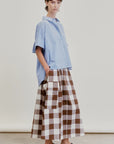 A person stands against a plain background wearing a loose light blue short-sleeve shirt paired with the Aquarius Cocktail Le Cargo Skirt—a long brown and white checkered cargo skirt crafted from Japanese cotton poplin, featuring oversized cargo pockets—and black woven slip-on shoes. Their hands are tucked into the pockets of the skirt.