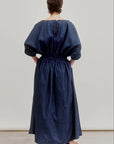 A person faces away, wearing the Aquarius Cocktail French 75 denim dress—loose-fitting, ankle-length with gathered sleeves, waist, and a boat neckline. They have short hair and flat sandals, set against a plain light background.
