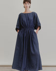 A person stands against a plain backdrop wearing Aquarius Cocktail's French 75, an ankle-length, loose-fit Japanese denim dress with a boat neck, elastic waist, short sleeves, and pockets; brown sandals complete the look.