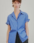 A person with short brown hair wears the Aquarius Cocktail Le Camp SS Shirt with Pocket, a blue Japanese cotton camp-style shirt featuring white pinstripes, paired with black pants against a plain light background.