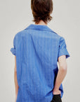 A person with short brown hair stands with their back to the camera, wearing the Aquarius Cocktail Le Camp SS Shirt with Pocket—blue Japanese cotton with thin white stripes—and black pants, one hand in their pocket, against a plain light background.