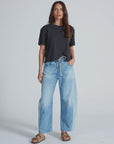 A woman with long brown hair and a relaxed expression stands against a plain background, wearing ASKK’s Cancun Pant—light blue Japanese denim jeans with wide legs and an elastic waistband—paired with a loose black t-shirt and tan sandals.