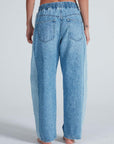 A person stands barefoot facing away, wearing light blue Cancun Pant jeans by ASKK with relaxed fit, back pockets, and an elastic waistband. The background is plain and light-colored.