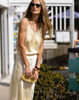 A woman in THE SHIRT's The Slip Skirt, sunglasses, and holding a yellow clutch stands outdoors by greenery and a white fence, sunlight highlighting her outfit.