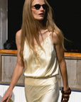 A woman with long blonde hair and black sunglasses walks outdoors, wearing THE SHIRT’s The Cami Shirt—a light yellow sleeveless top with a No Gape button—paired with a yellow clutch and chunky bracelet.