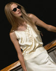 A woman with long blonde hair wears black sunglasses and The Cami Shirt by THE SHIRT, featuring a No Gape button, standing by a wooden railing with a dark background.