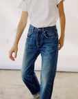 A person wears Hey Gang's "The Loose Crop" t-shirt, paired with relaxed, mid-rise jeans made of 100% cotton denim on a concrete floor. Their face is not visible, their hand rests relaxed by their side, and the backdrop is plain white.