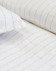 Close-up of white bed sheets and pillowcases featuring the Blake Big Pillow 28x36 from POM POM, showcasing light gray interwoven stripes. The fabric appears soft and neatly arranged, creating a clean and minimalist look.