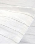 A close-up of the Blake Big Pillow 28x36 by POM POM reveals a white fabric adorned with subtle, sophisticated gray stripes, highlighting its hand-loomed texture and pattern. The material appears soft and lightweight, elegantly folded at an angle.
