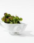 The Montes Doggett Gravy Boat No. 151, a whimsical, white high-fired ceramic container with short legs and a handle, is filled with green chive blossoms. Handmade in Peru, the container casts a soft shadow on the clean, white background.