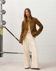 A woman with long, straight hair stands confidently in a studio, wearing the CLOSED Jayla Denim jeans, a brown suede jacket, and a beige top. Industrial equipment can be seen in the background.