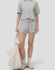A woman wears the CLOSED Cropped Rugby Shirt in light gray with matching drawstring shorts, white sneakers, socks, and an off-white tote bag. The look features a relaxed silhouette against a plain, light-colored background.