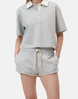 A woman wears the CLOSED Cropped Rugby Shirt in light gray with a white collar and buttons, paired with matching drawstring shorts, standing hands-in-pockets against a plain background.